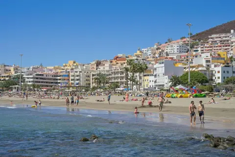 Getty Images Locals and tourists swim, walk and play on a sandy beach. The water is clear and calm and there is a backdrop of palm trees and colourful apartment buildings in the background