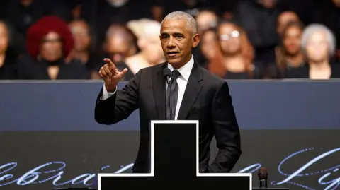 Getty Images Former US president Barack Obama speaks at the memorial service, wearing a black suit and tie. He's standing on stage with a microphone in front of him and a large cross donning the podium.
