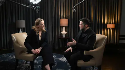 Laura Kuenssberg (L) sits talking to Ukrainian President Volodymyr Zelensky. Kuenssberg wears a black suit jacket, black trousers and cream top. Her hands are clasped beside her hip. Zelensky sits oposite her and is wearing a black suit jacket, black shirt and black trousers. His hand is slightly raised as he talks. The pair are sat on beige chairs, behind them is a grey curtain and three lamps. The carpet beneath them is dark blue with a cream floral pattern.