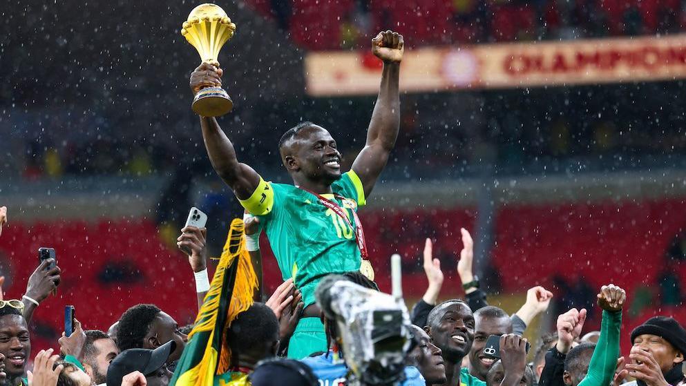 Senegal player Sadio Mane, dressed in green kit and wearing a golden medal on a red ribbon around his neck, is held aloft by fellow players as he raises both arms into the air, holding the Africa Cup of Nations trophy in his right hand.